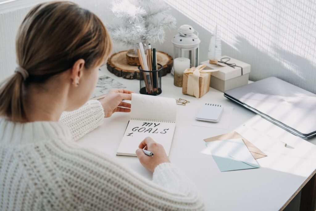 A woman writing her goals in a desk with other table stuff.